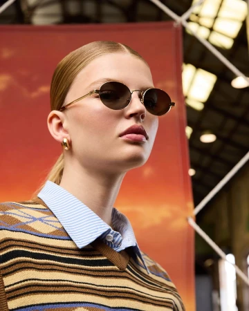 A fair-skinned adult woman with blond hair and round sunglasses poses indoors against a warm-toned backdrop wearing a striped sweater.