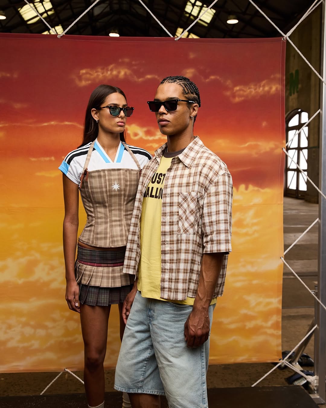 Two people model sunglasses in front of sunset backdrop. Woman in miniskirt and man in plaid shirt in an indoor industrial location.