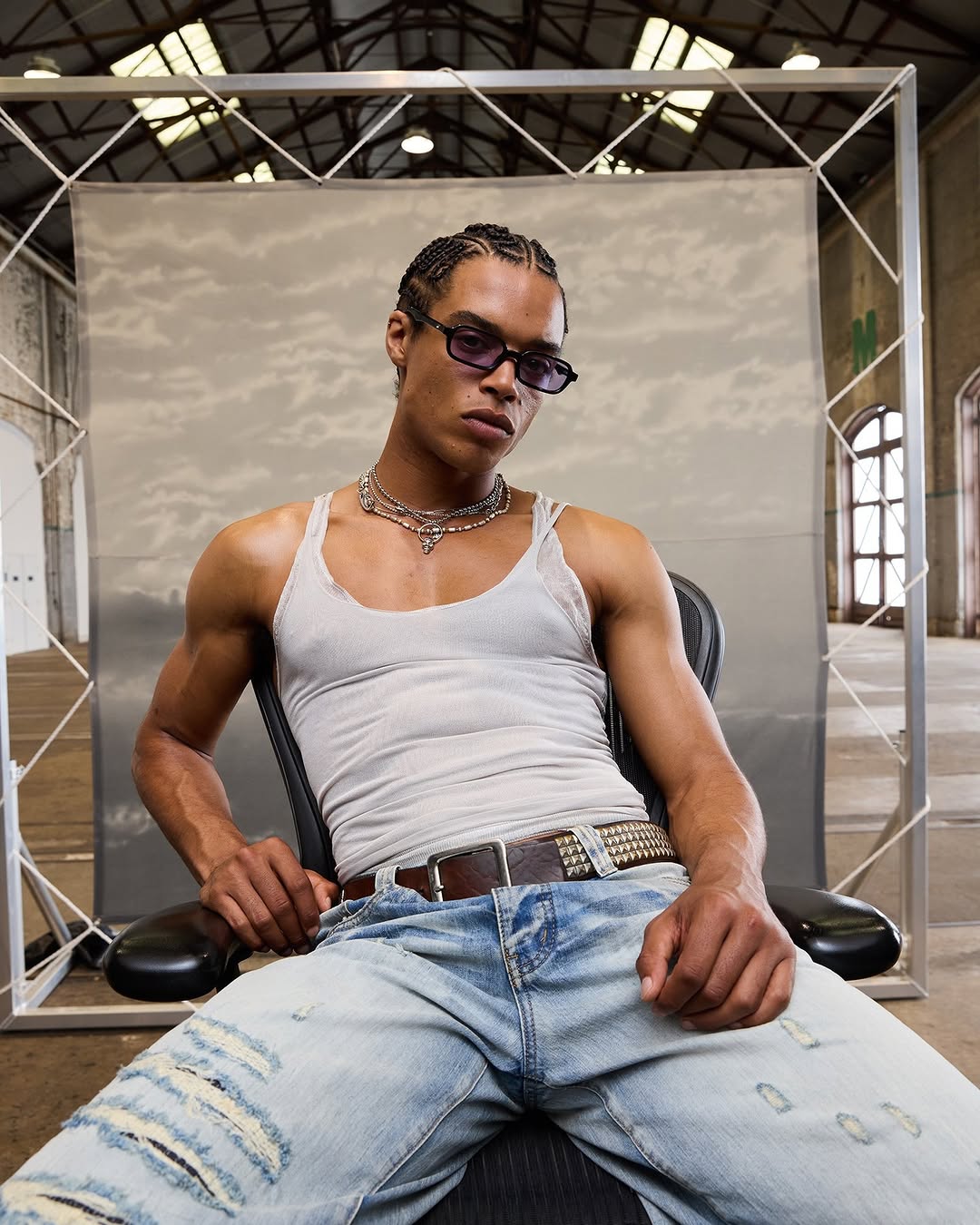 Portrait of a young adult male with braided hair, wearing sunglasses, a white tank top and ripped blue jeans, sitting in a chair indoors.