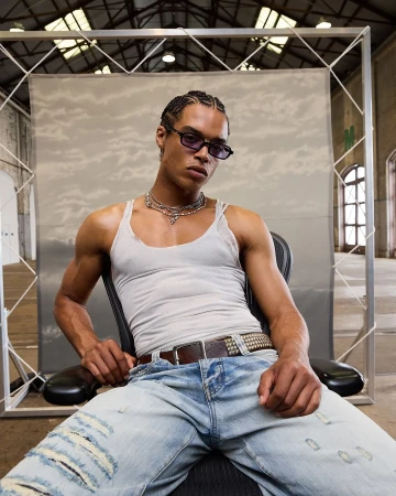 Portrait of a young adult male with braided hair, wearing sunglasses, a white tank top and ripped blue jeans, sitting in a chair indoors.