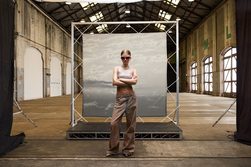 A young adult woman stands with arms crossed in front of a grey backdrop in a large indoor space with a high ceiling and arch windows.