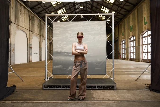 A young adult woman stands with arms crossed in front of a grey backdrop in a large indoor space with a high ceiling and arch windows.