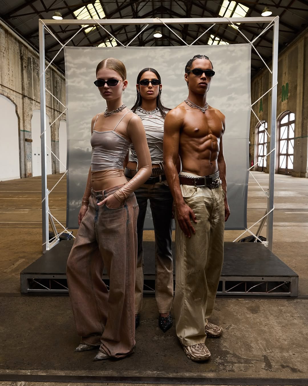 Three fashion models wearing sunglasses, neutral tops and baggy pants pose in a warehouse with a backdrop and industrial architecture.