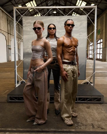 Three fashion models wearing sunglasses, neutral tops and baggy pants pose in a warehouse with a backdrop and industrial architecture.