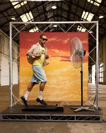 Man walks on treadmill in industrial setting with fan and backdrop. He wears sunglasses, yellow t-shirt, plaid shirt, and casual shorts.