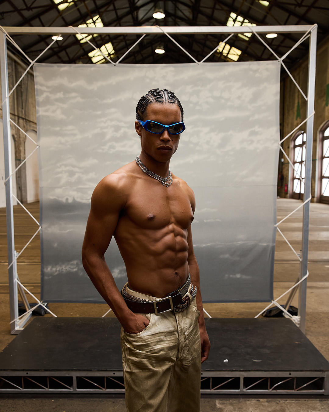 Shirtless male model with cornrows, wearing blue sunglasses and a silver chain, poses in front of a cloudy backdrop in a warehouse.