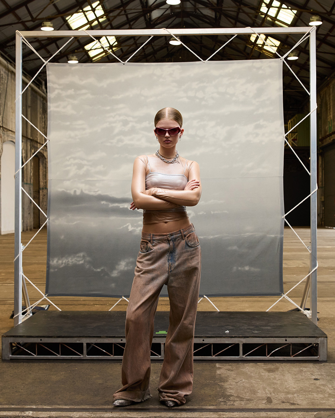 Young woman poses on a platform in baggy jeans, sunglasses, and top against a cloud-print backdrop inside an industrial space.