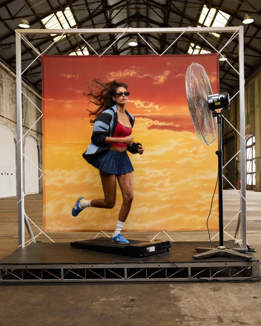 A brunette woman in athletic wear runs on a treadmill with a sunset backdrop and industrial fan in a large warehouse-like space.
