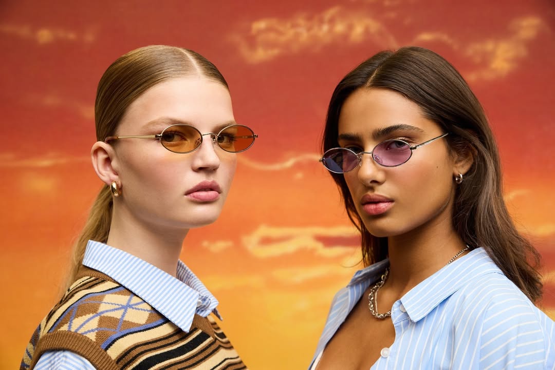 Two women pose in front of an orange sunset backdrop wearing oval sunglasses and trendy outfits in a close-up portrait.