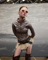 Model wearing oval sunglasses, metallic jacket, and pleated skirt posing in front of a printed cloud backdrop