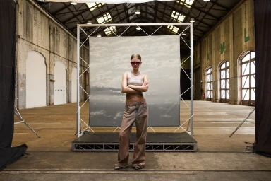 Model wearing sunglasses and baggy jeans posing in front of a framed cloud backdrop inside a large warehouse