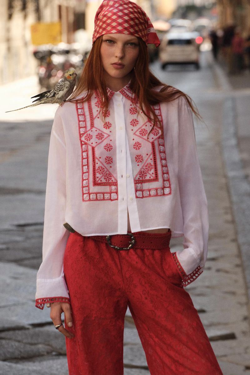 Fashionable red-haired woman wearing a white collared shirt with red embroidery, red pants, and a matching bandana stands outdoors.