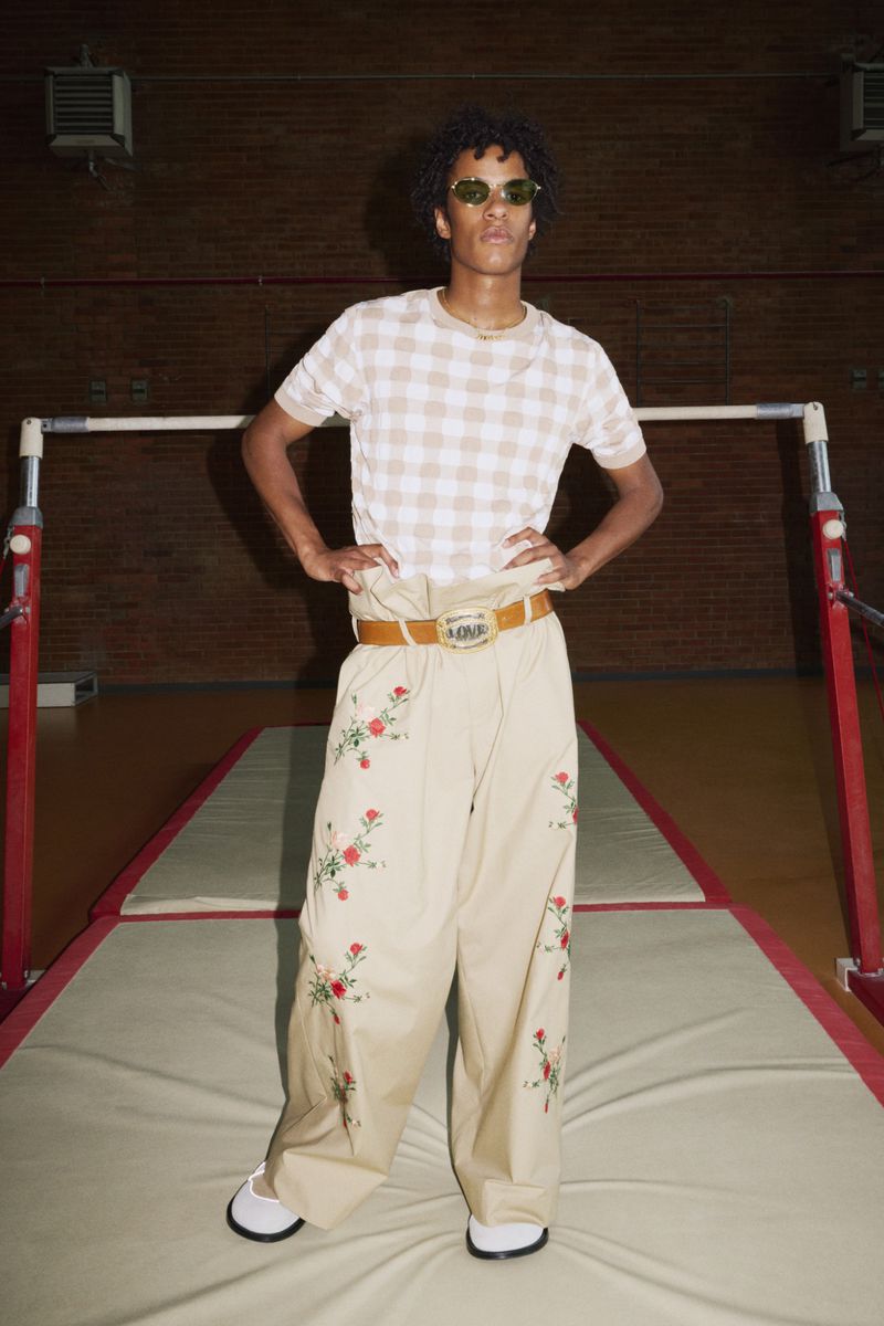 Young adult with curly hair poses in a beige and white checkerboard top and embroidered trousers in a gymnasium setting.