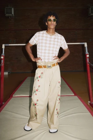 Young adult with curly hair poses in a beige and white checkerboard top and embroidered trousers in a gymnasium setting.