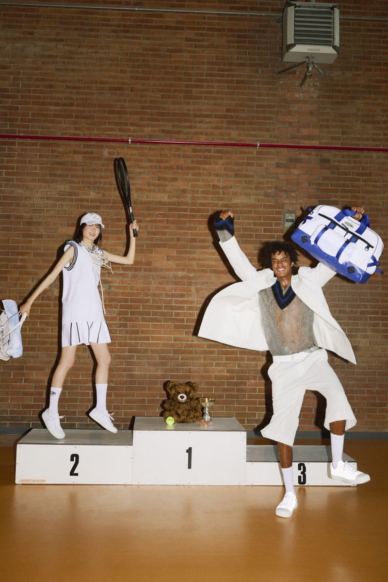Two athletes stand on a numbered podium with props, in an indoor court setting with a brick wall in the background.