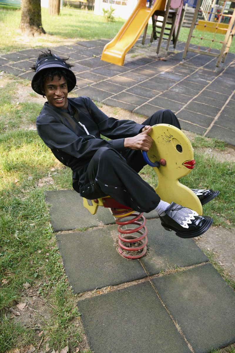 A stylish adult rides a yellow duck spring rider on a playground, smiling. He wears a black outfit and bucket hat, and patterned socks.