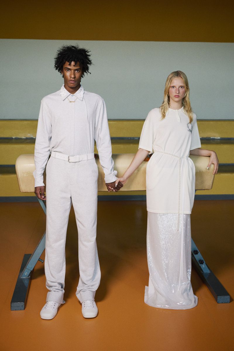 Two models in cream-colored clothing hold hands in a gymnasium, standing in front of a vintage vaulting horse with a yellow wall backdrop.