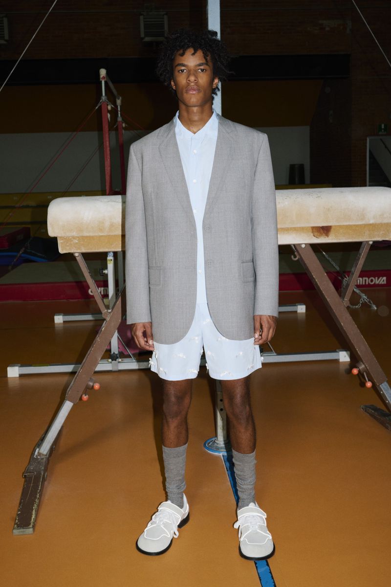 A young adult man stands in a gymnasium wearing a blazer, shirt, and shorts, posing next to a gymnastics vault.