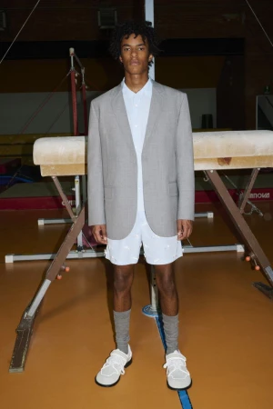 A young adult man stands in a gymnasium wearing a blazer, shirt, and shorts, posing next to a gymnastics vault.
