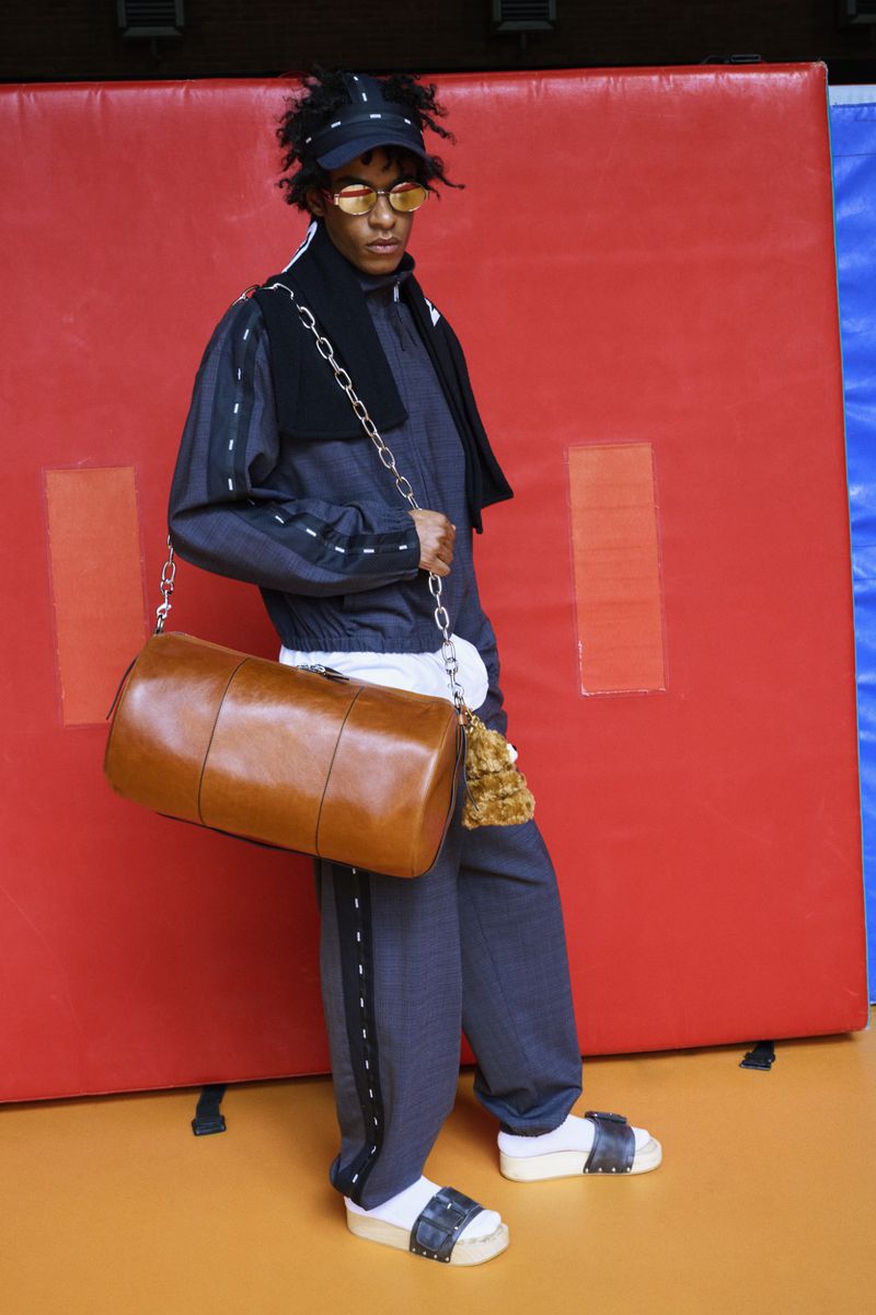 Full shot of a person in a navy tracksuit with white trim, sunglasses, and sandals, carrying a brown leather duffel bag in a studio setting.