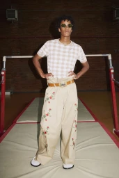 Model wearing a plaid t-shirt, tan embroidered trousers, and sunglasses standing in an indoor sports gym.