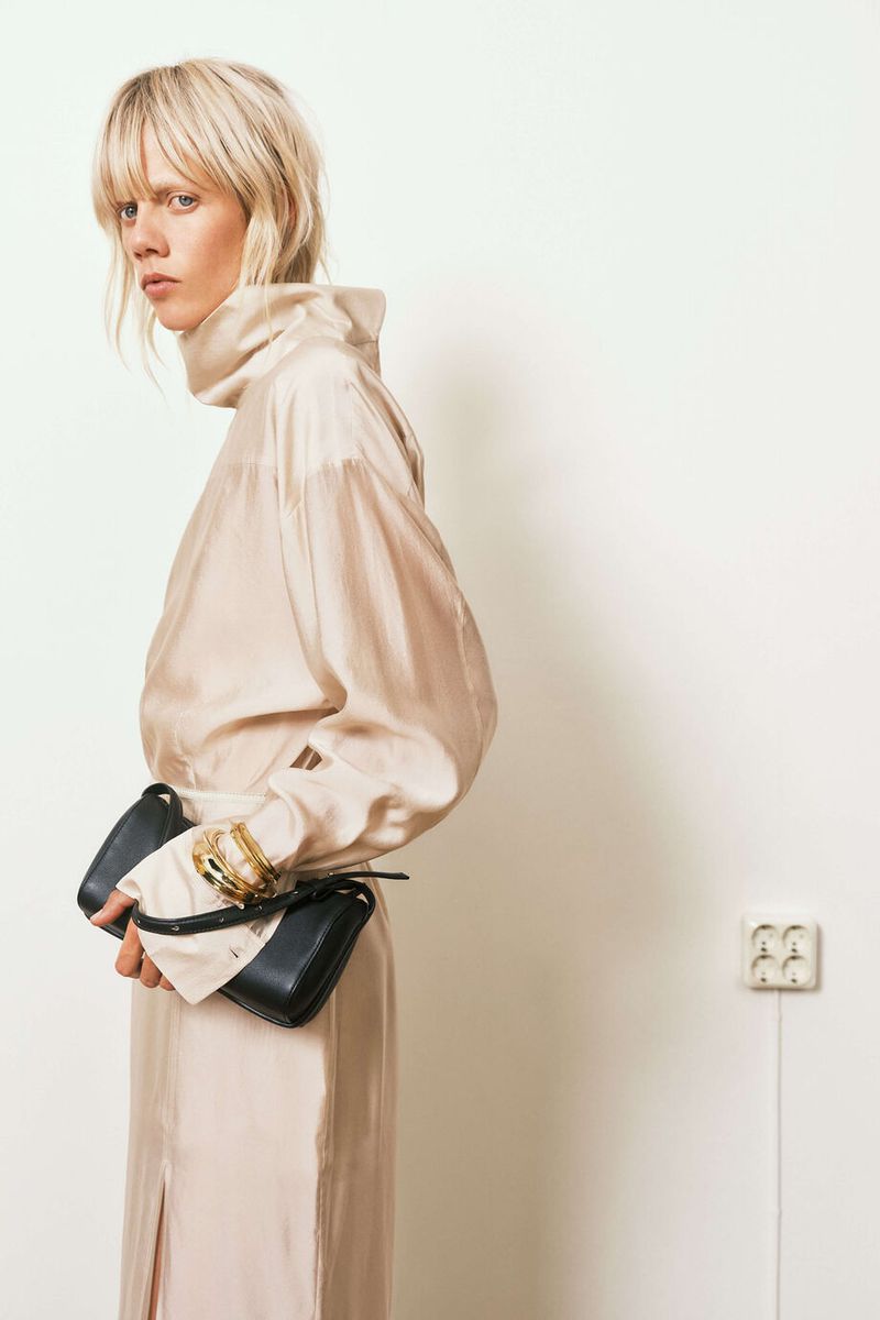 Stylish woman in a pale blouse and long skirt holds a black handbag in an indoor setting with a plain white background.