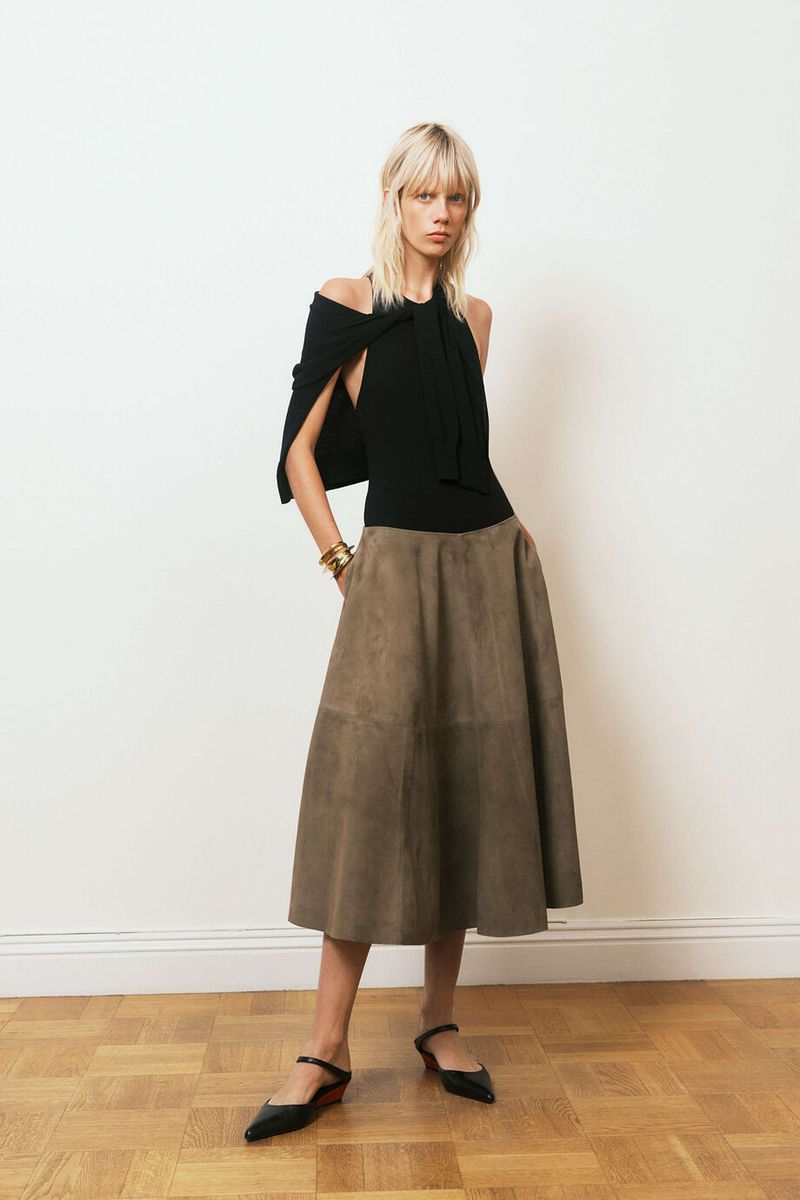 A fair-skinned adult woman poses in a studio wearing a black top and khaki skirt, complemented by black shoes and gold bracelets.