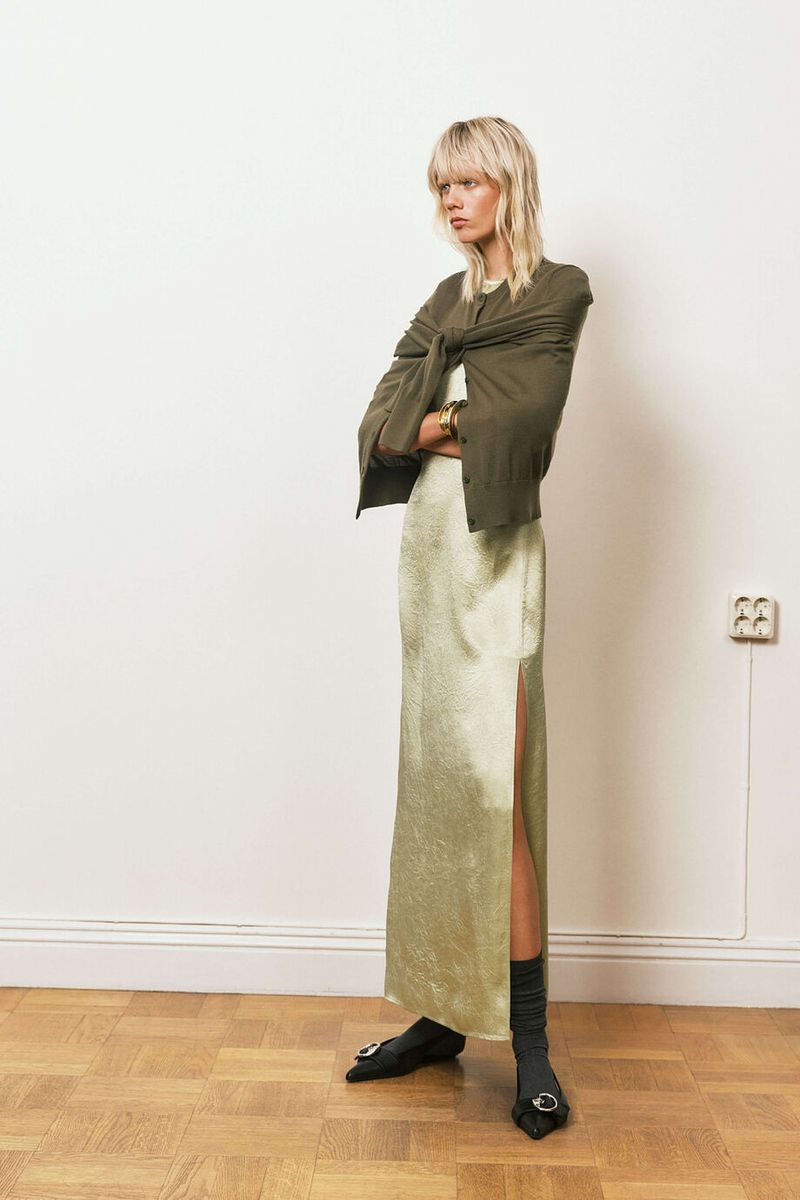 Full-length shot of a female fashion model in a green dress with a thigh-high slit, a sweater tied around her shoulders, and black flats.