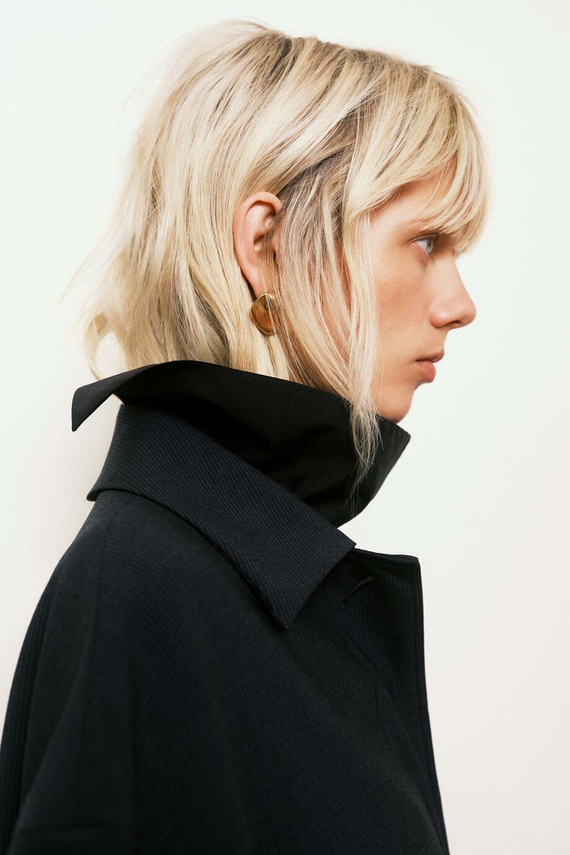 Profile shot of a fair-skinned adult female with wavy blonde hair, wearing a dark collared coat in front of a neutral background.