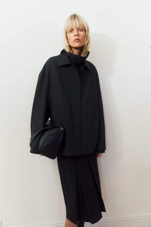 Model in a black jacket and matching skirt holding a dark handbag against a plain white wall for Filippa K.