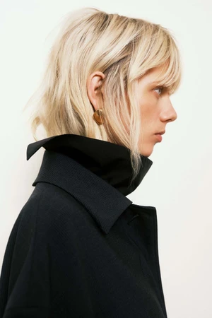 Profile portrait of a blonde model wearing a dark collared coat and an earring against a white background