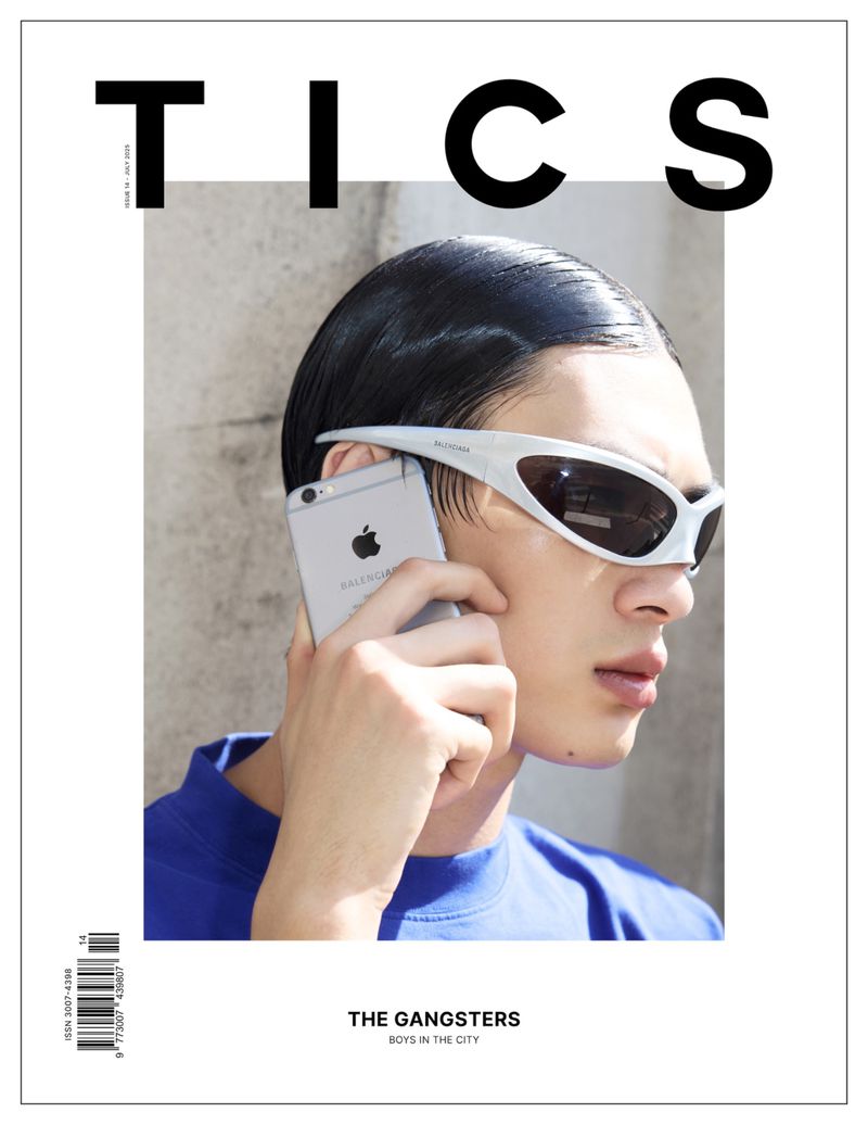 Fashion magazine cover featuring a young adult male wearing white sunglasses, talking on a silver smartphone with a blurred grey background.