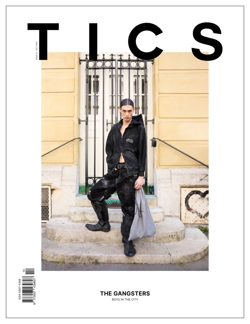 Magazine cover shot of a young adult male in black clothing standing in front of a building with stone steps, holding a grey bag.
