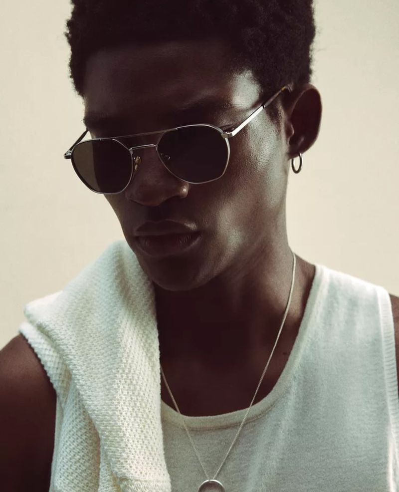 Portrait of an adult man wearing silver-framed aviator sunglasses, a white tank top, and a silver pendant necklace against a neutral background.