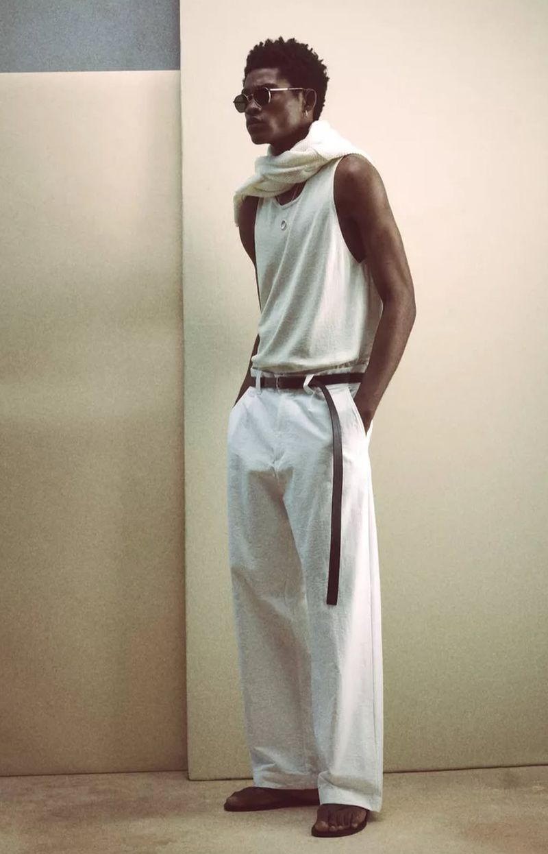Fashionable African American man posing in a tank top and white wide-leg pants, accessorized with a scarf and sunglasses, against a neutral backdrop.