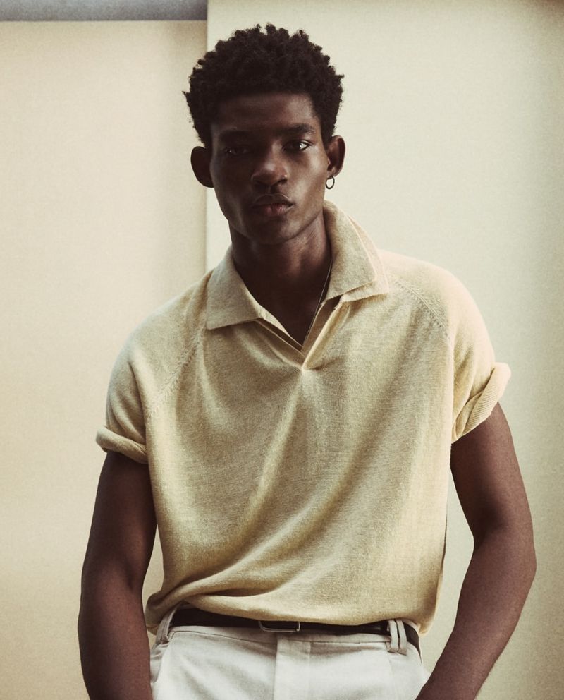 Portrait of a dark-skinned male model wearing a beige polo shirt and white pants, posing in front of a neutral wall indoors.