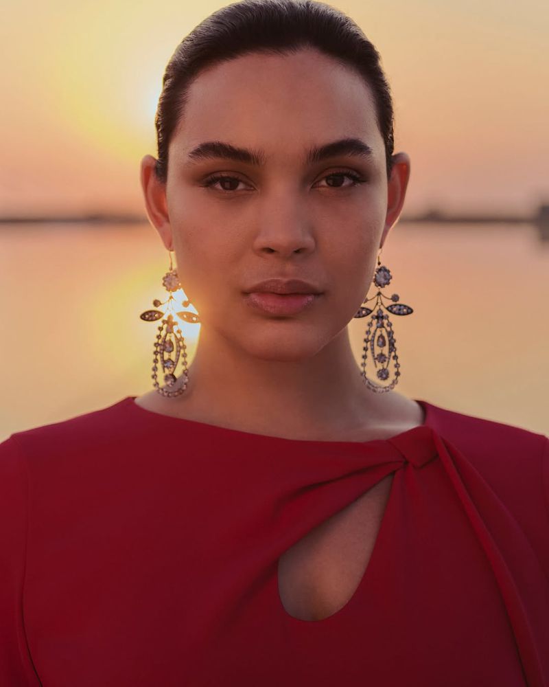 Portrait of a woman with elegant earrings in a red dress, set against a warm-toned lake backdrop during golden hour.