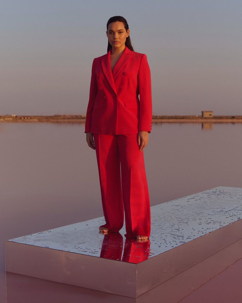 Elegant adult woman in a red pantsuit stands on a platform in a pink salt lake, set against a soft blue sky at sunset.
