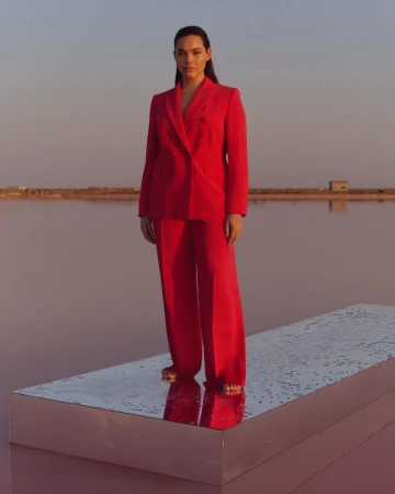 Elegant adult woman in a red pantsuit stands on a platform in a pink salt lake, set against a soft blue sky at sunset.