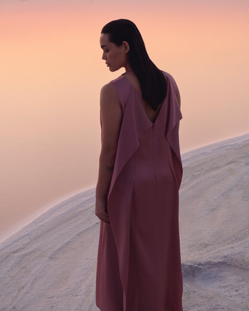 A person in a long rose colored dress stands outdoors on white ground with a soft gradient sky. The person gazes to the side.