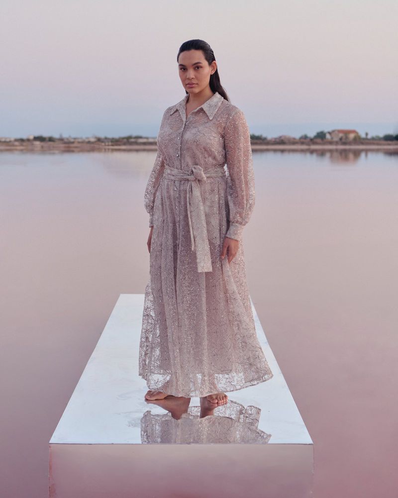 Woman in a long-sleeved sheer dress with a belt stands on a reflective platform in a pink lake at sunset, looking at the camera.