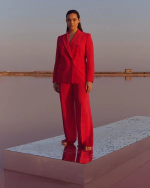 Model wearing a vibrant red trouser suit standing barefoot on a reflective mirrored platform overlooking a calm body of water at sunset.