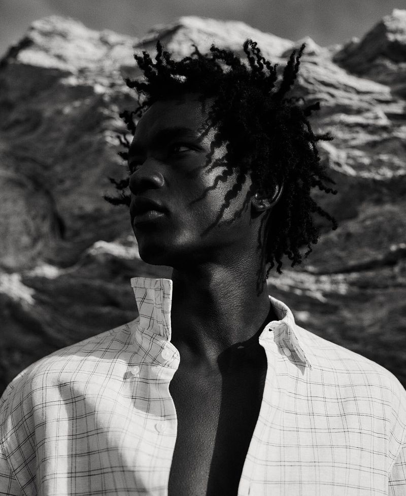 Monochrome portrait of a young adult male with dreadlocks wearing a patterned white dress shirt against a rocky background.