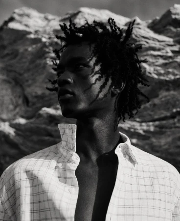 Monochrome portrait of a young adult male with dreadlocks wearing a patterned white dress shirt against a rocky background.
