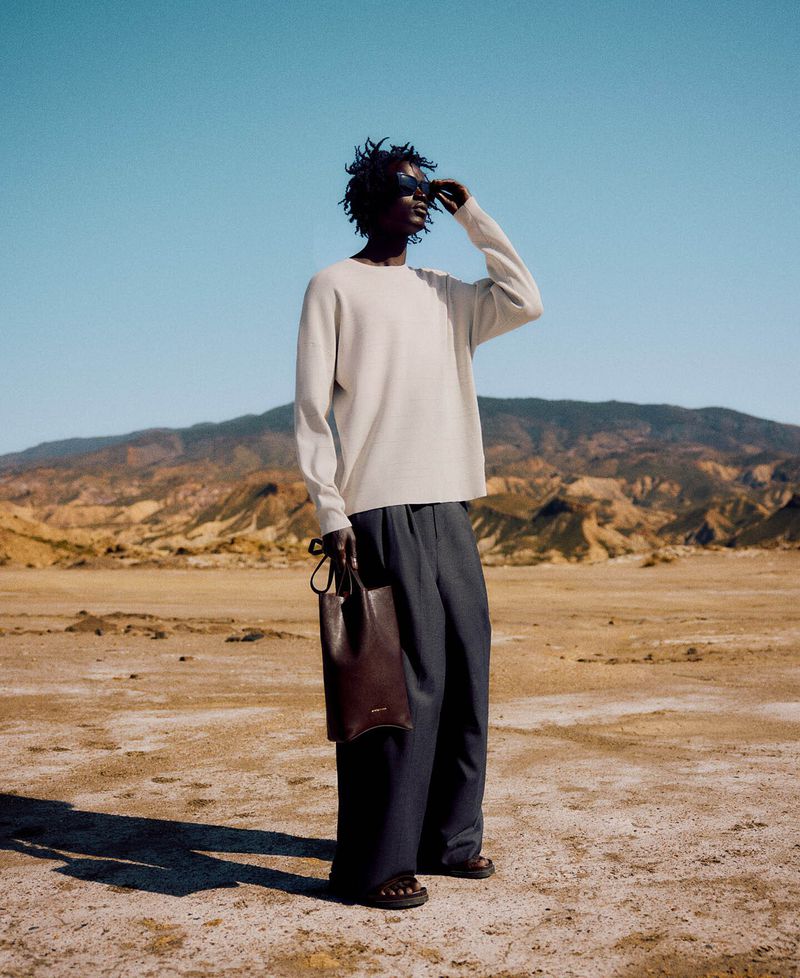 An adult male in sunglasses shields his eyes in a desert landscape wearing a long-sleeve shirt, trousers, and sandals with a leather bag.