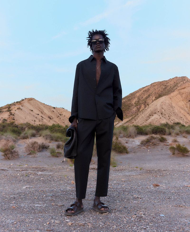 Man in unbuttoned black shirt and pants stands outside with leather bag in hand, in front of desert hills under a light sky.