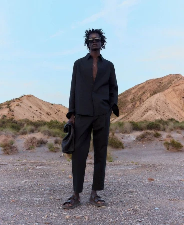 Man in unbuttoned black shirt and pants stands outside with leather bag in hand, in front of desert hills under a light sky.