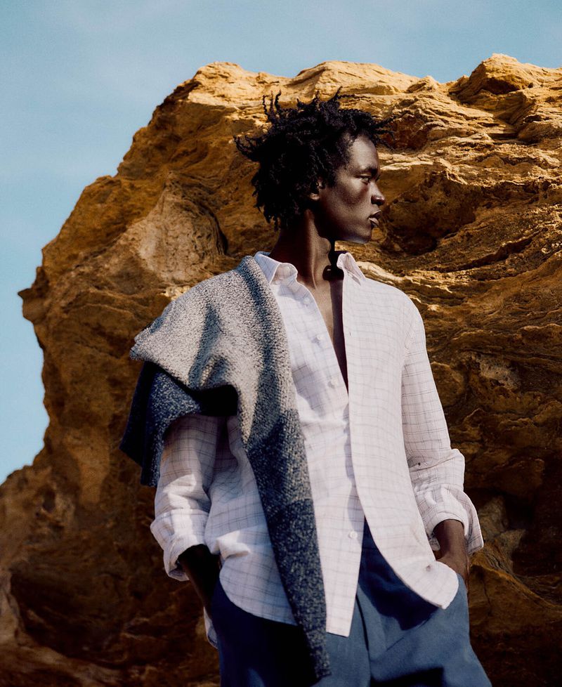Portrait of a black male fashion model outdoors. He wears a dress shirt and sweater with a rock formation and sky in background.