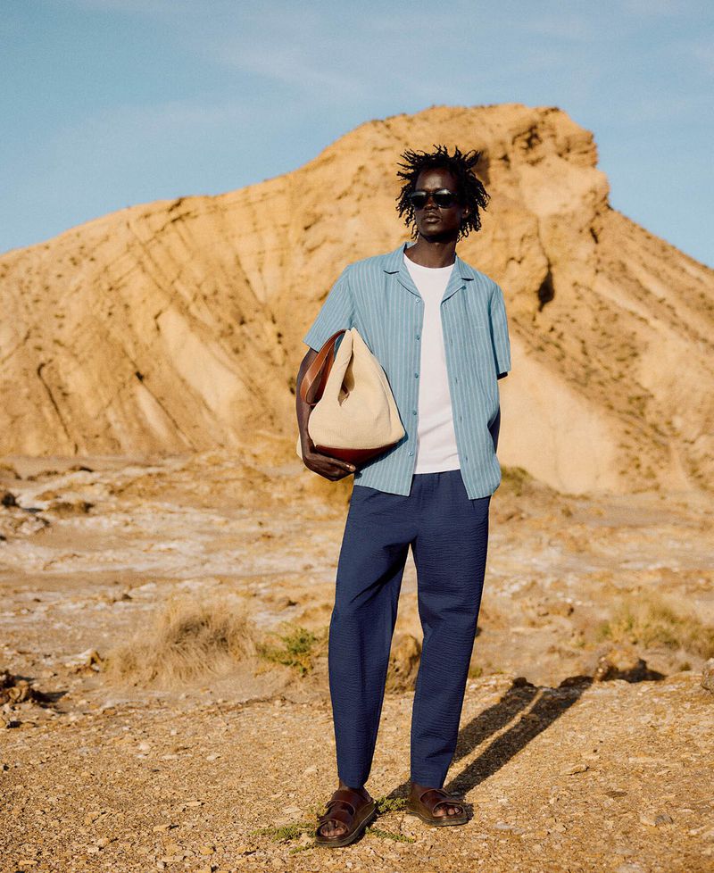 Adult male in casual resort wear holding tote bag in desert setting with clear blue sky, showcasing fashion and style.