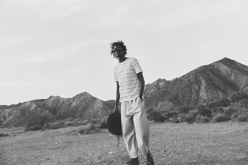 Monochrome portrait of a man in casual wear holding a hat in a desert landscape with mountains in the background, looking at the camera.